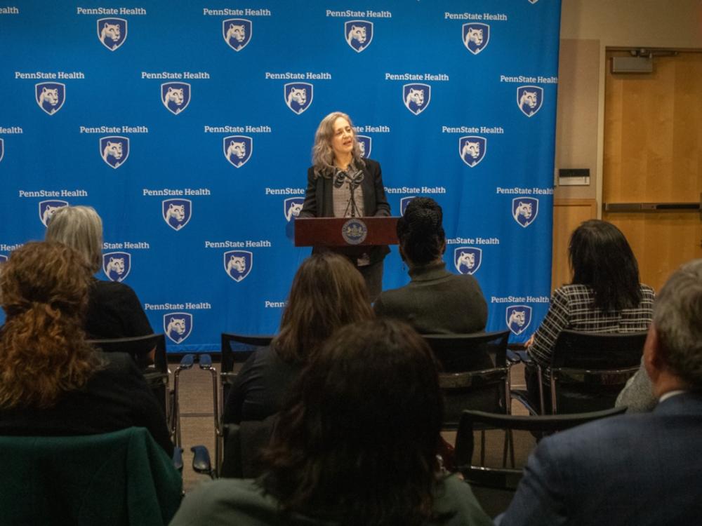 A woman in professional attire speaks at a lectern, gesturing with her hands. A step-and-repeat ‘Penn State Health’ backdrop is in the background.