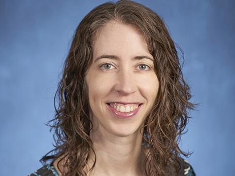 Erica Frankenberg head shot with long, wavy brown hair, black, blue, and green print short-sleeved blouse, smiling.