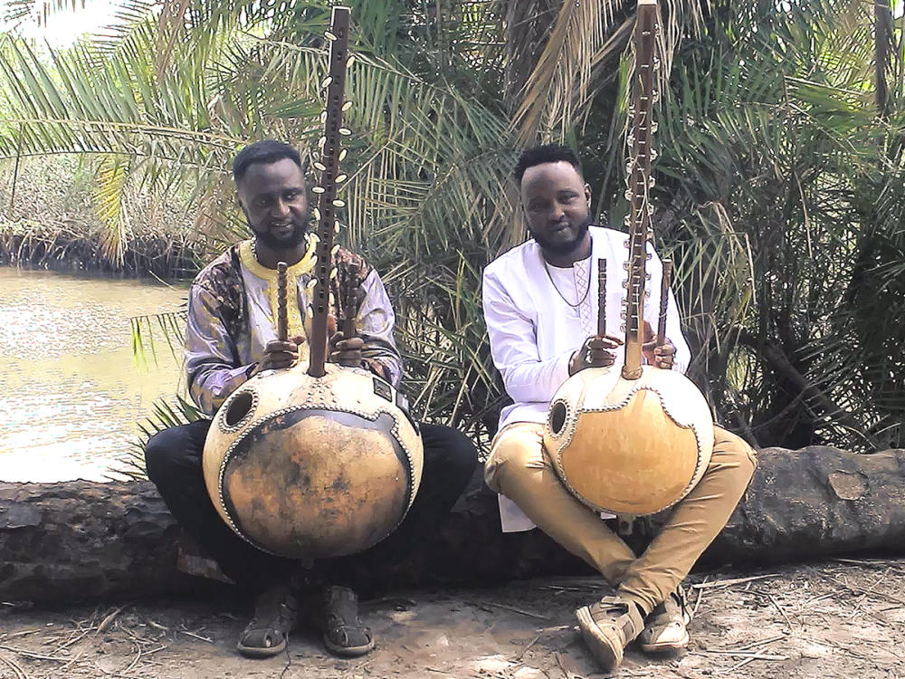 Two musicians pose outside near a river with their instruments
