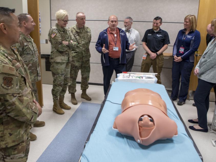 A group of National Guard members in uniform and medical professionals in civilian attire stand around a hospital bed featuring a medical training mannequin. One person in a jacket appears to be explaining or demonstrating something, while the others listen attentively in a clinical setting.