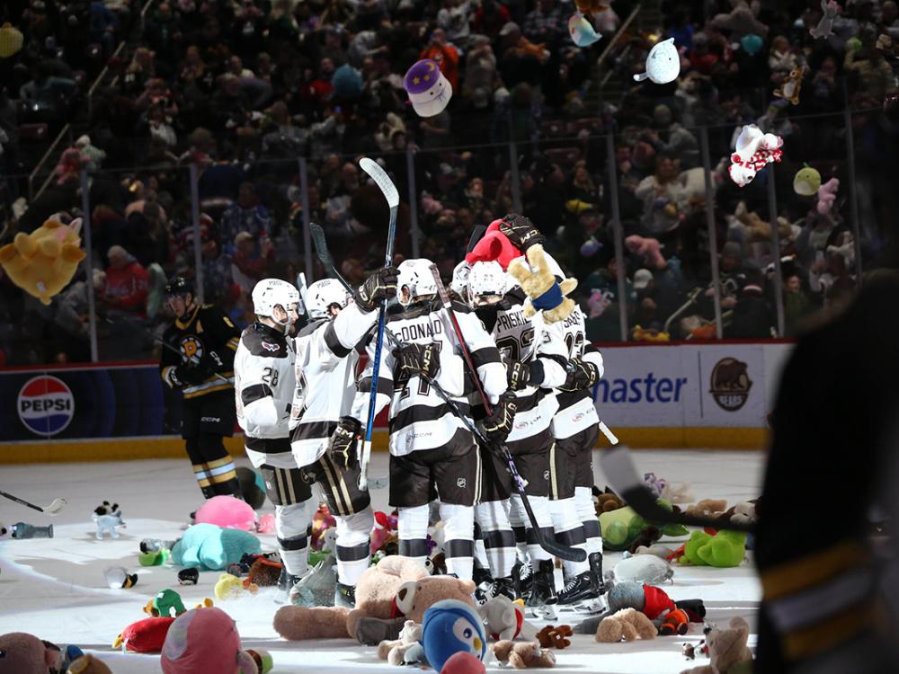 A group of professional hockey players gather facing each other on a hockey rink. Stuffed animals are on the ice around them and flying through the air from the spectators who are throwing them.