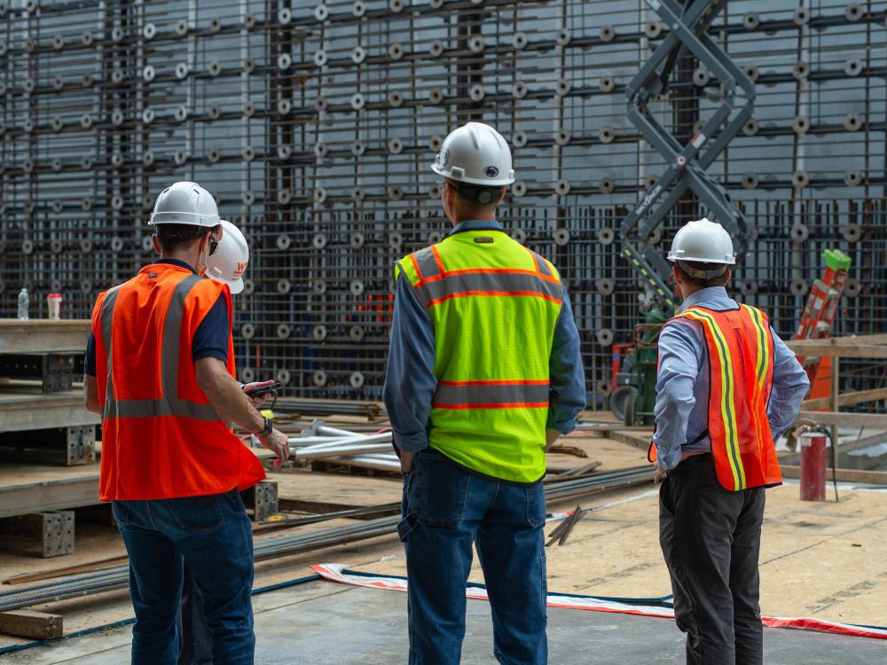 Four people in hard hats and neon safety vests converse while standing in a construction zone