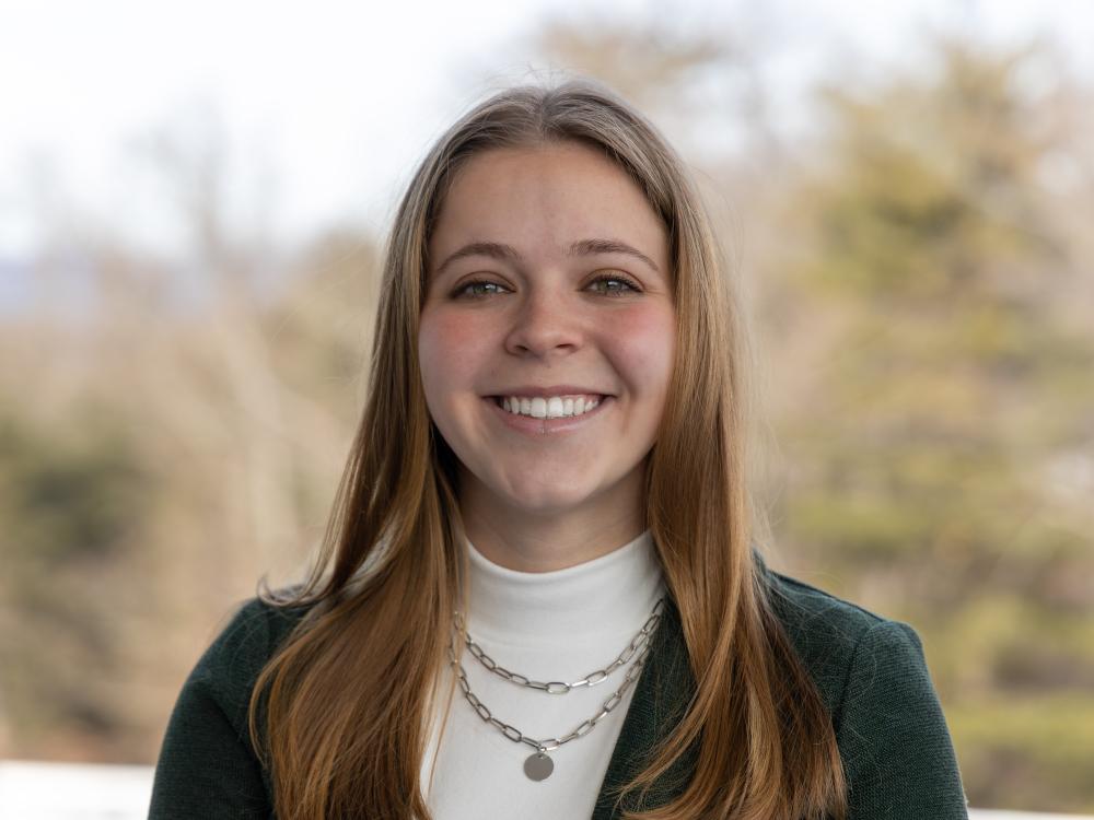 Cara Arnoldi smiles in a green and white sweater in front of a blurred background of green and brown trees.