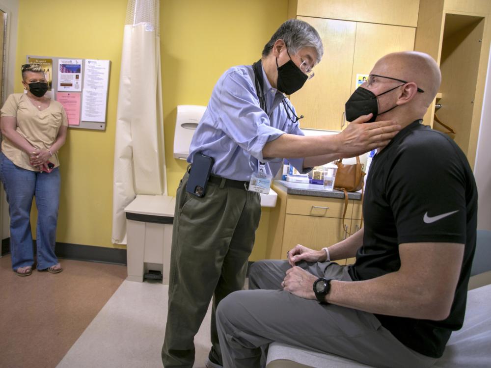 Shin Mineishi, center, of the Penn State Cancer Institute, touches the neck of a patient, Doug Harris, in a patient exam room. On the left, Harris’ wife Lisa stands and watches. All three are wearing face masks.