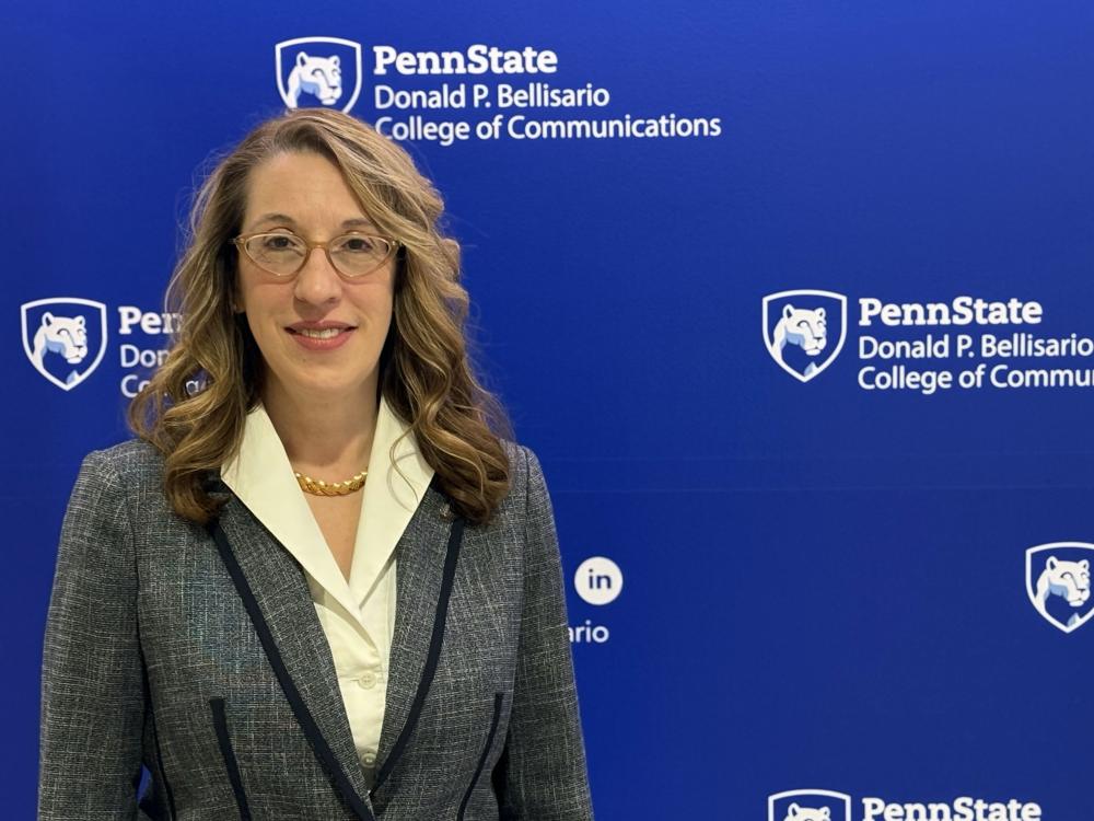 A woman with brown hair and glasses wearing a suit standing in front of a blue wall. 