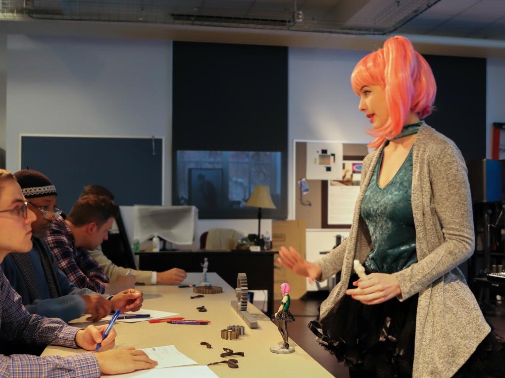 A student presents her 3d model figurine concept to a panel of judges during the Arts Business Idea Competition. Mimicking the look of her 3d model, she wears a pink ponytail wig and vibrant outfit.