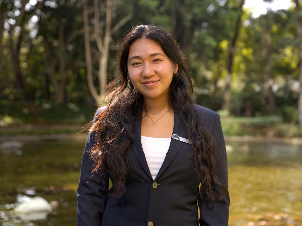 Jamie Nguyen smiles in a navy blazer standing in front of a pond and trees.