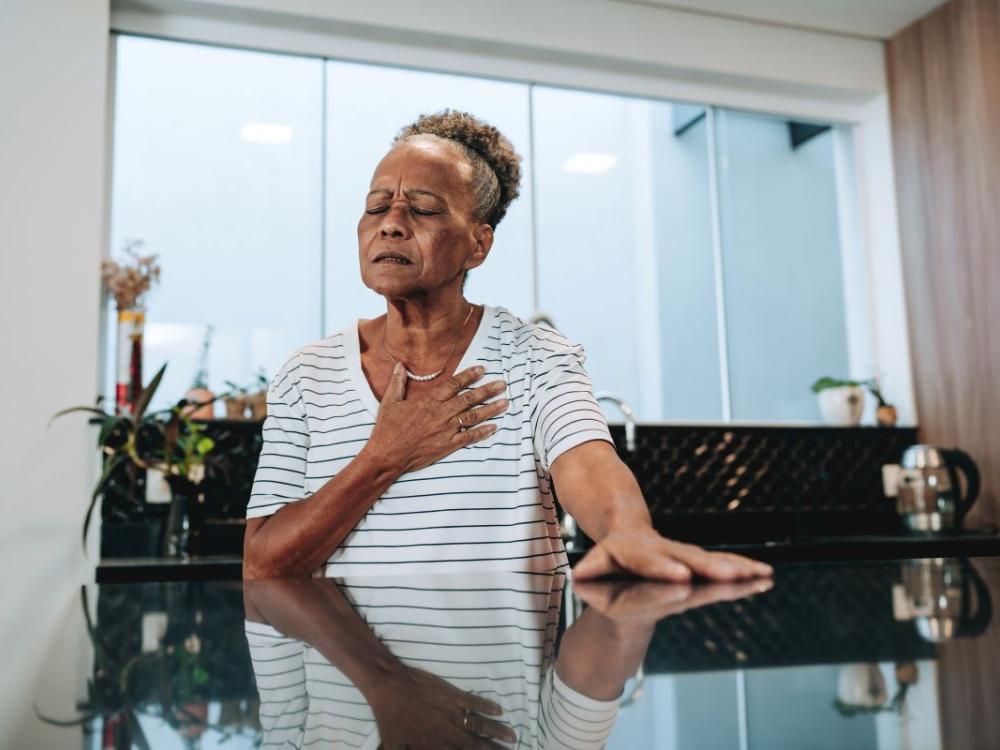 An elderly woman puts her hand over in chest and grimaces in pain.