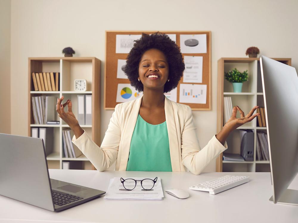 A Black woman sits at a desk, smiling and holding her hands up to either side.