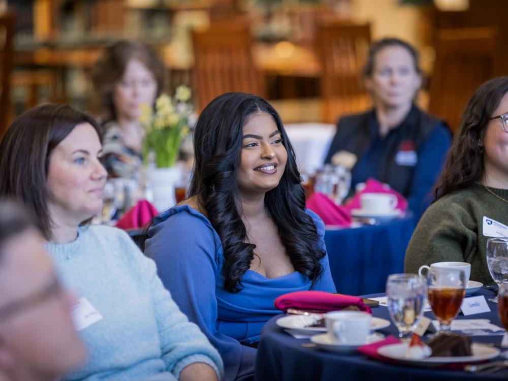A student sits with a group of women at a luncheon program.