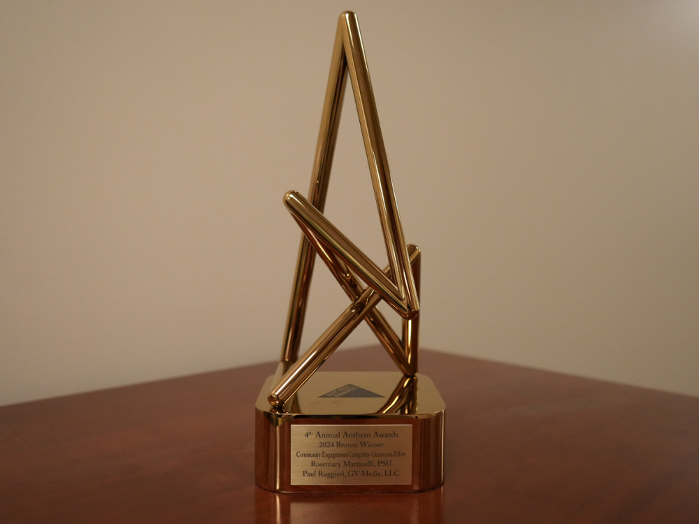 A bronze anthem award on a table, the words "4th Annual Anthem Awards 2024 Bronze Winner; Community Engagement Categories-Grassroots Effort; Rosemary Martinelli, PSU; Paul Ruggieri, GV Media, LLC" are engraved upon its base.