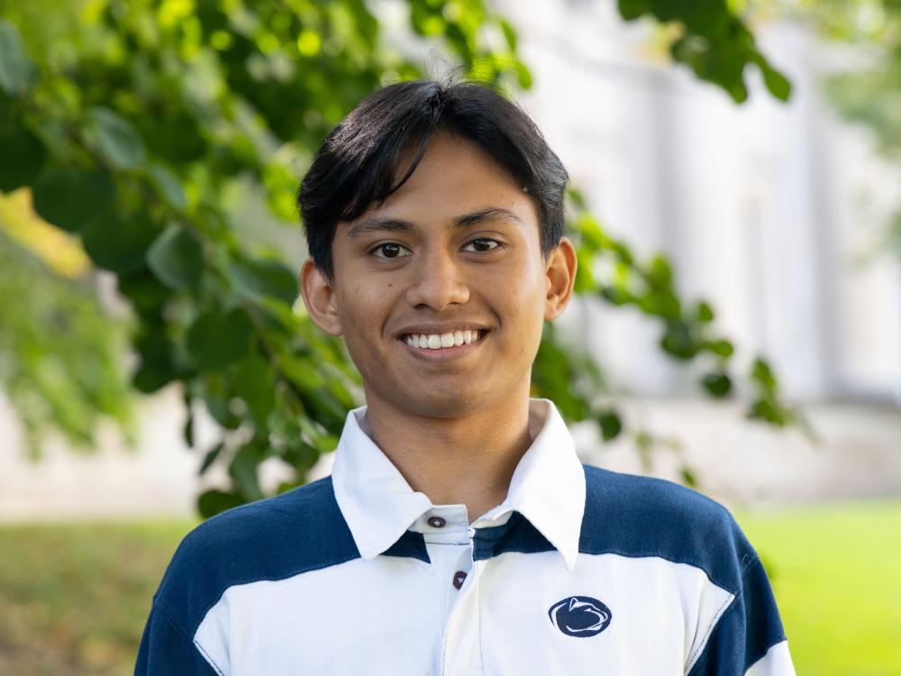 Muhammad Nazaruddin bin Mohd Salleh smiles while wearing a blue and white Penn State shirt in front of a tree outside Sparks Building.