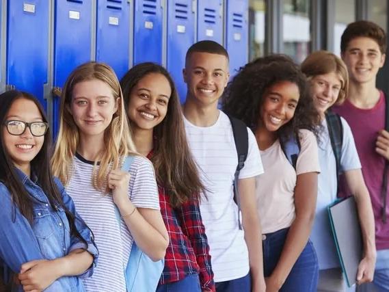 A group of teens stand smiling at the camera against a background of lockers