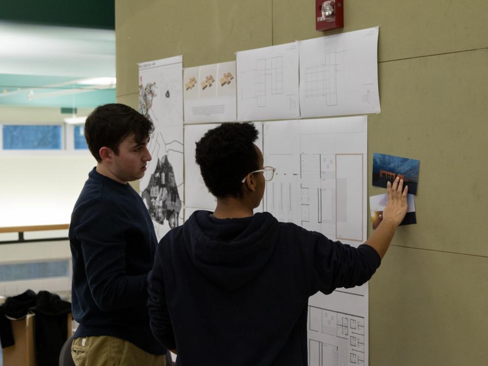 Two students look at architectural designs tacked to a wall.