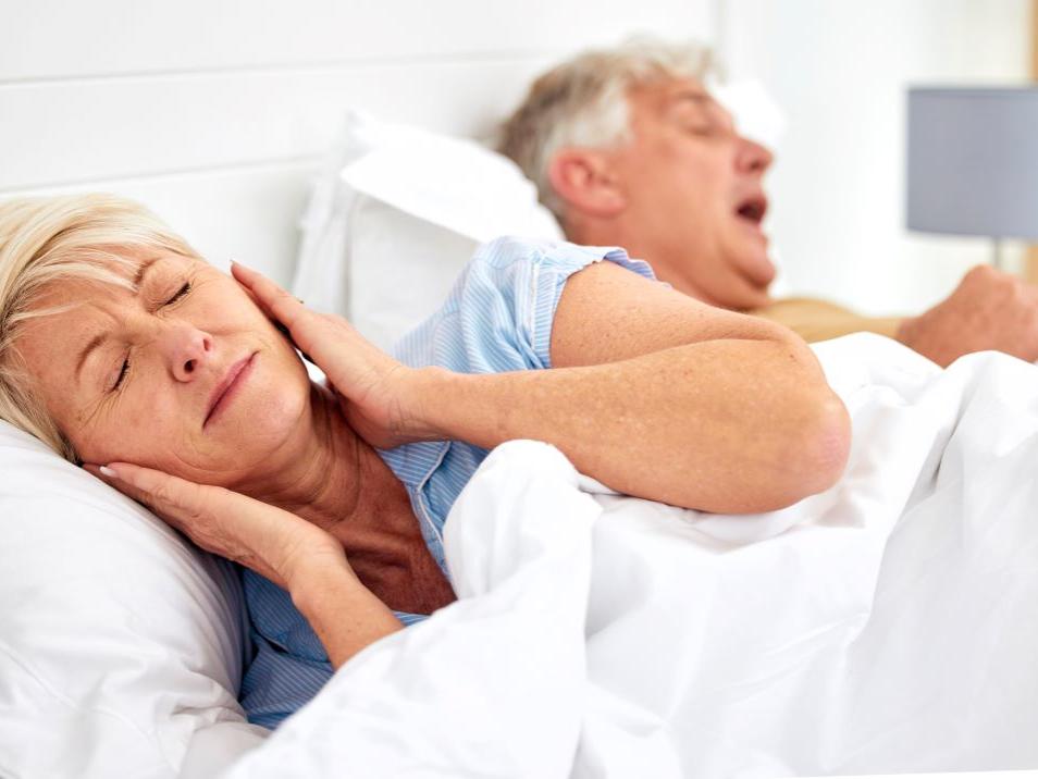 An older woman has her hands over her ears as her husband lies next to her snoring.
