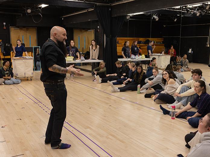Man in a black shirt and pants speaks in front of a group of students gathered around him in a circle