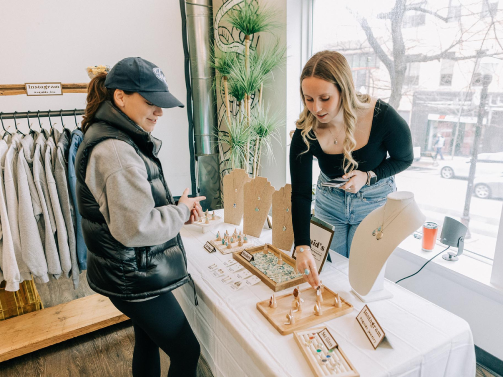 One student stands behind a table selling jewelry on display, another student stands in front of the table looking at the displayed jewelry.