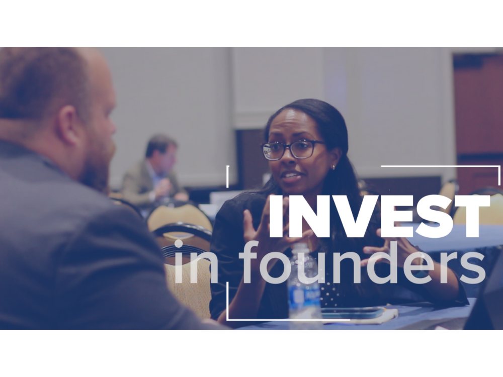 The words "Invest in Founders" appears overtop of an image of an individual sitting at a table explaining their business to an investor