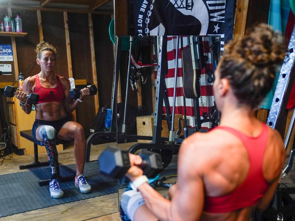 Woman wearing workout clothing lifts weights while sitting on a bench. She has a prosthetic leg.
