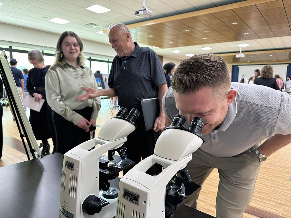 A man looks through a microscope while a student talks to another man