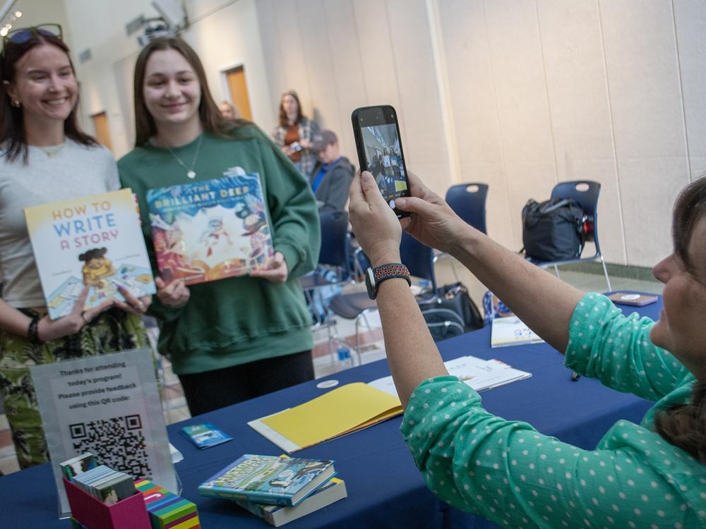 Two students pose holding children's books, while a woman takes a photo with her phone