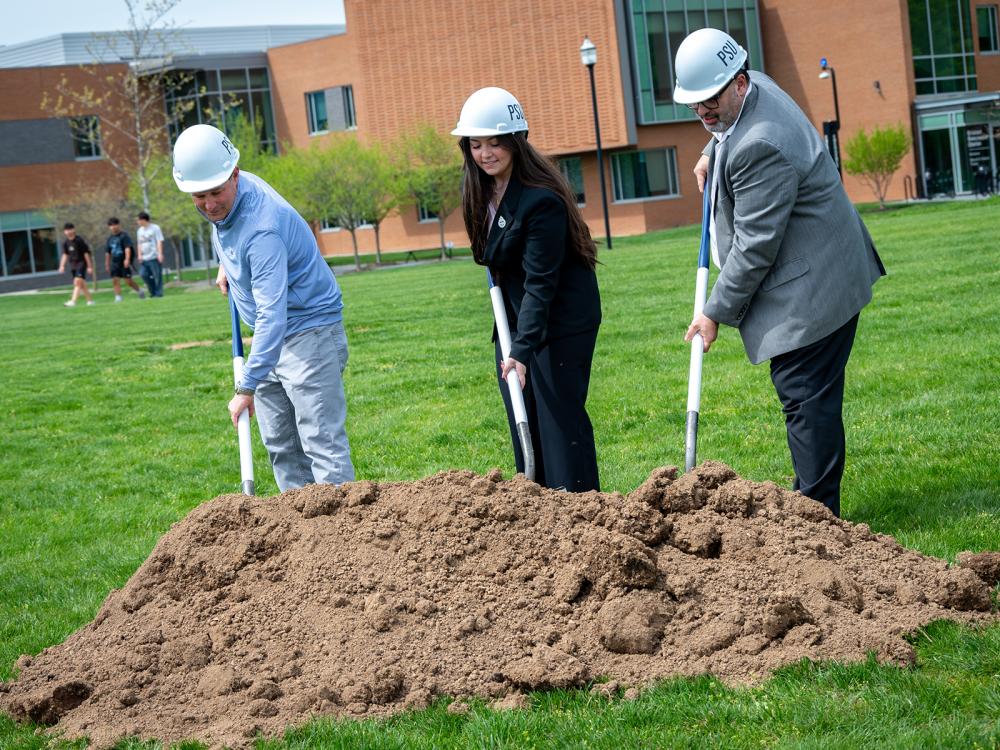 Three people wearing hard hats push shovels into a pile of dirt