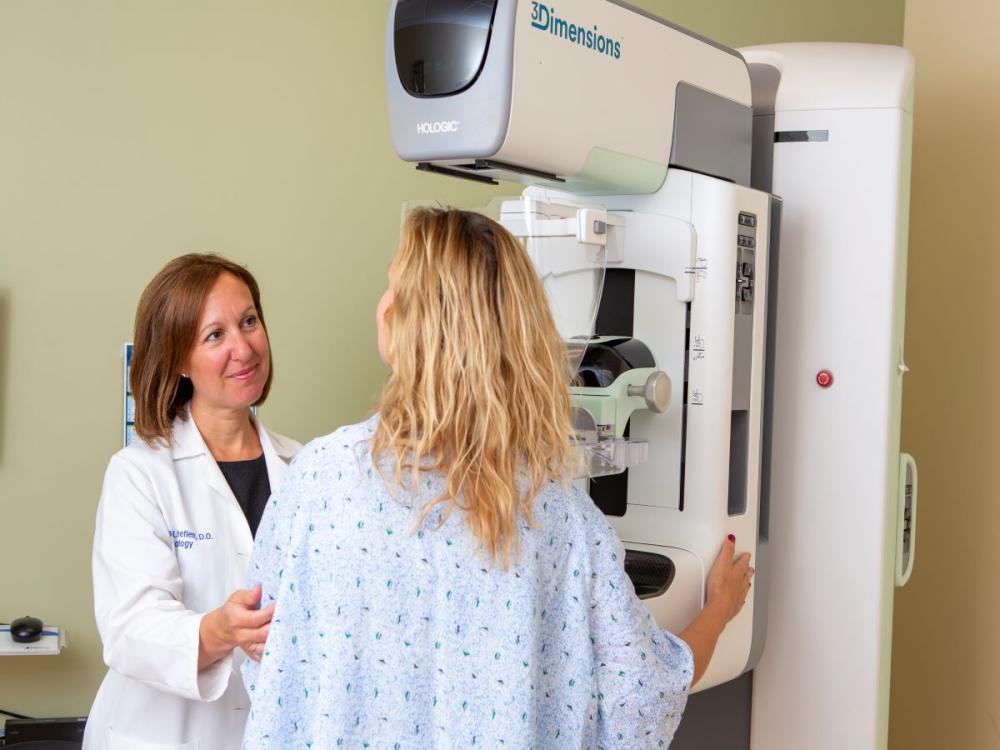Alison Chetlen, left, touches a patient’s arm as she positions her in front of a mammography machine. Chetlen is wearing a white coat with her name and “Radiology” on the upper-right.