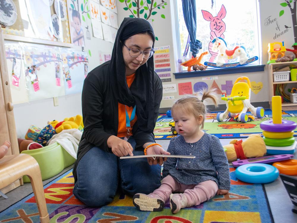 A woman reads a book to a young child in a day care center, surrounded by colorful toys