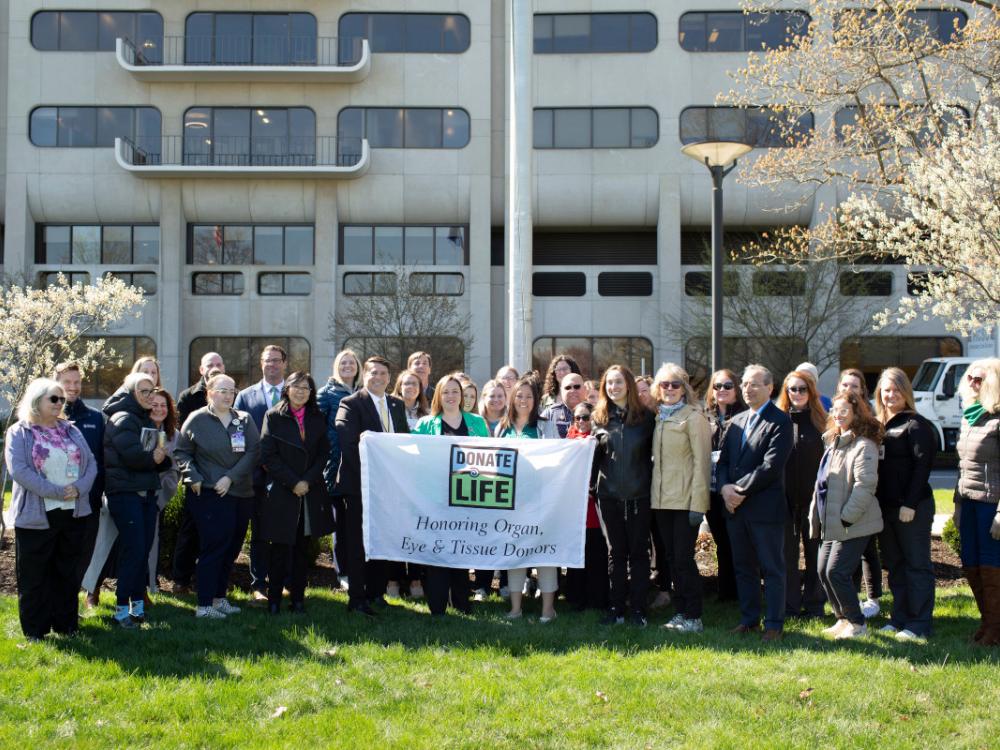 A group of people pose with a “Donate Life” flag. A hospital building with windows is in the background.