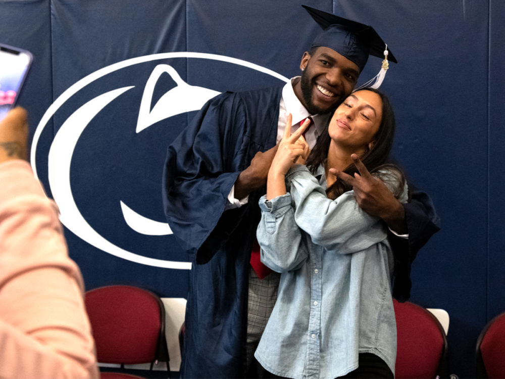 A graduate in cap and gown poses with a woman flashing peace signs as they smile for a photo in front of a Penn State backdrop.