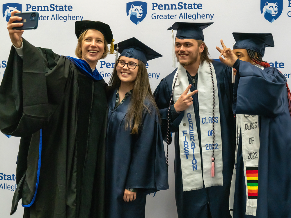 Megan L. Nagel in academic regalia takes a selfie with three graduates in caps and gowns, all smiling and posing in front of a Penn State Greater Allegheny step-and-repeat backdrop.