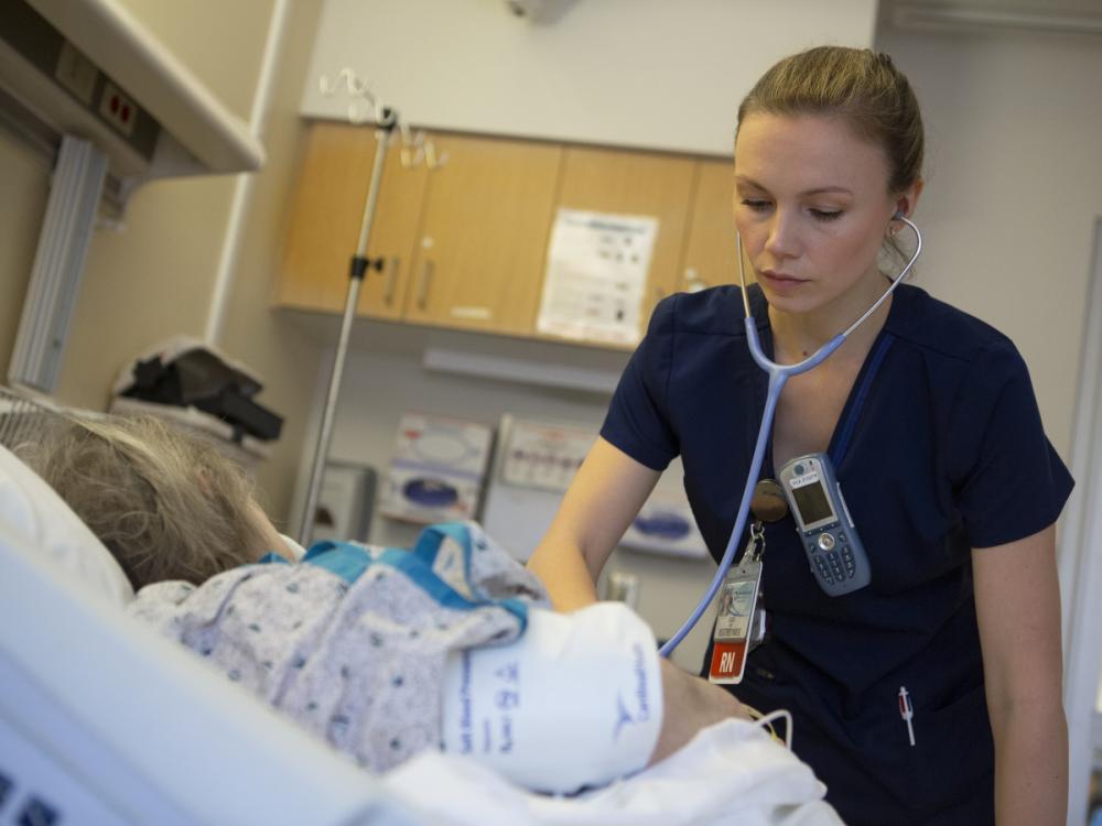 A nurse uses a stethoscope as she tends to a young patient in a hospital bed. The nurse also has a phone and a nametag clipped to her shirt. An IV pole and wooden cabinets are in the background.