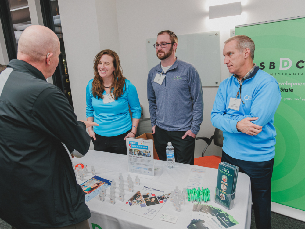 Three individuals stand behind a table with SBDC materials, speaking to an individual on other side of table.