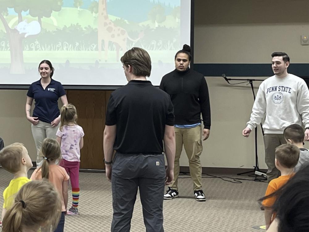 Students standing in front of a projector screen speaking demonstrating for preschool children