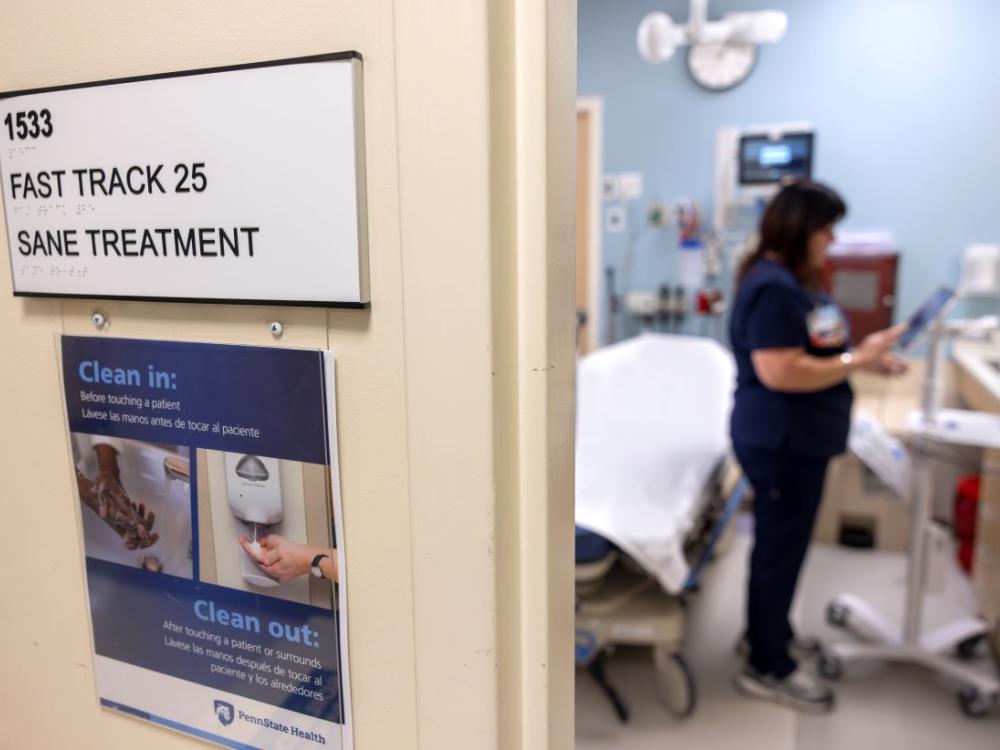 A nurse is seen out of focus on the right in the Sexual Assault Nurse Examiner room at Penn State Health St. Joseph Medical Center, A sign on the left reads: “Fast Track 25, SANE Treatment.” Another sign reads: “Clean in, Clean out.”