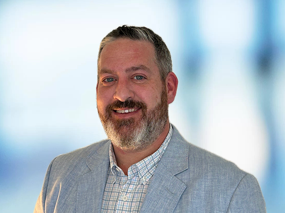 A headshot of a man with a beard wearing a light blue dress jacket