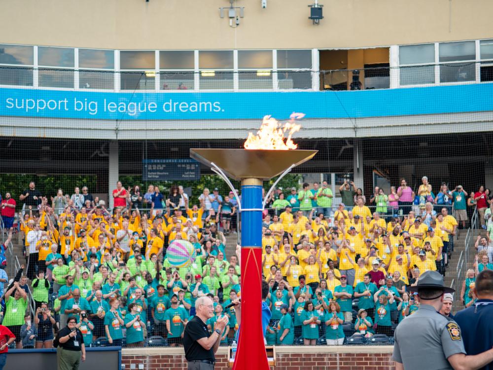 A huge lit torch is backgrounded by stadium stands full of people wearing colorful t-shirts