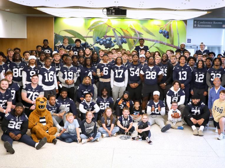 A large group photo of the Penn State football team, all wearing navy blue jerseys, posing with children and staff in a brightly lit hospital lobby.