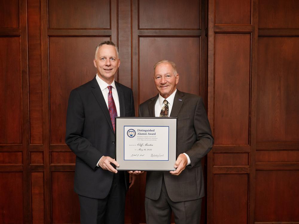 Two people stand together, one holds a framed award