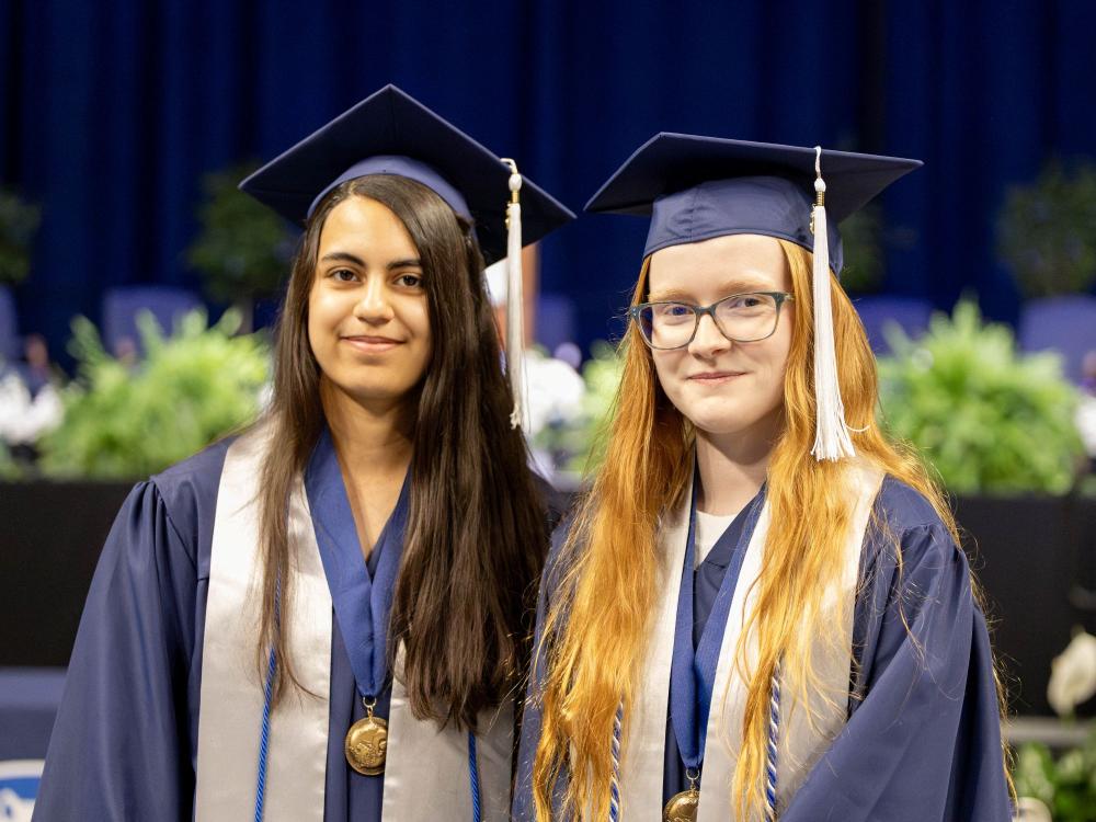 Fiona Combs (left) and Sé Morrisson (right)—two spring 2025 quadruple-major graduates—stand next to each other in front of the stage at the College of the Liberal Arts commencement ceremony in the Bryce Jordan Center on May 10, 2025.