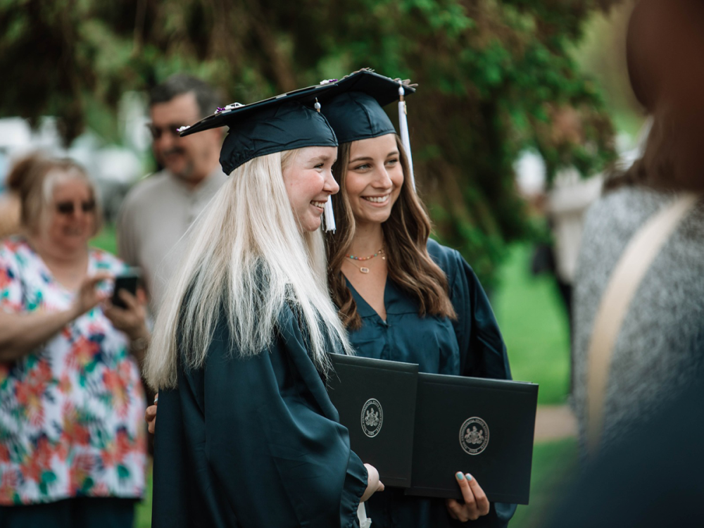 : Two graduates in caps and gowns smile while holding their diplomas during as friends and family take photos in the background.