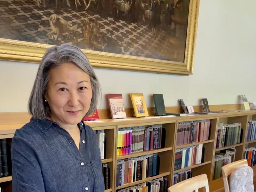Author, Julie Park, wearing a blue denim shirt and holding a large book open in a room with bookshelves