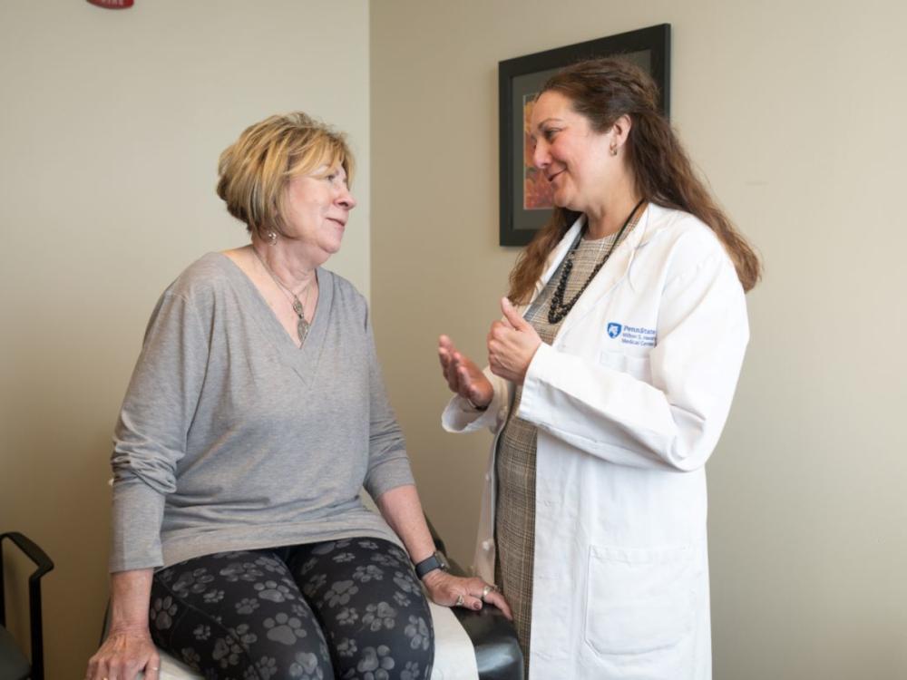 A woman sits on an exam table talking to Jaime Long, right, of Penn State Health. Long is gesturing with her hands and wearing a white coat with the Penn State Health Milton S. Hershey Medical Center logo on it. Both are smiling.