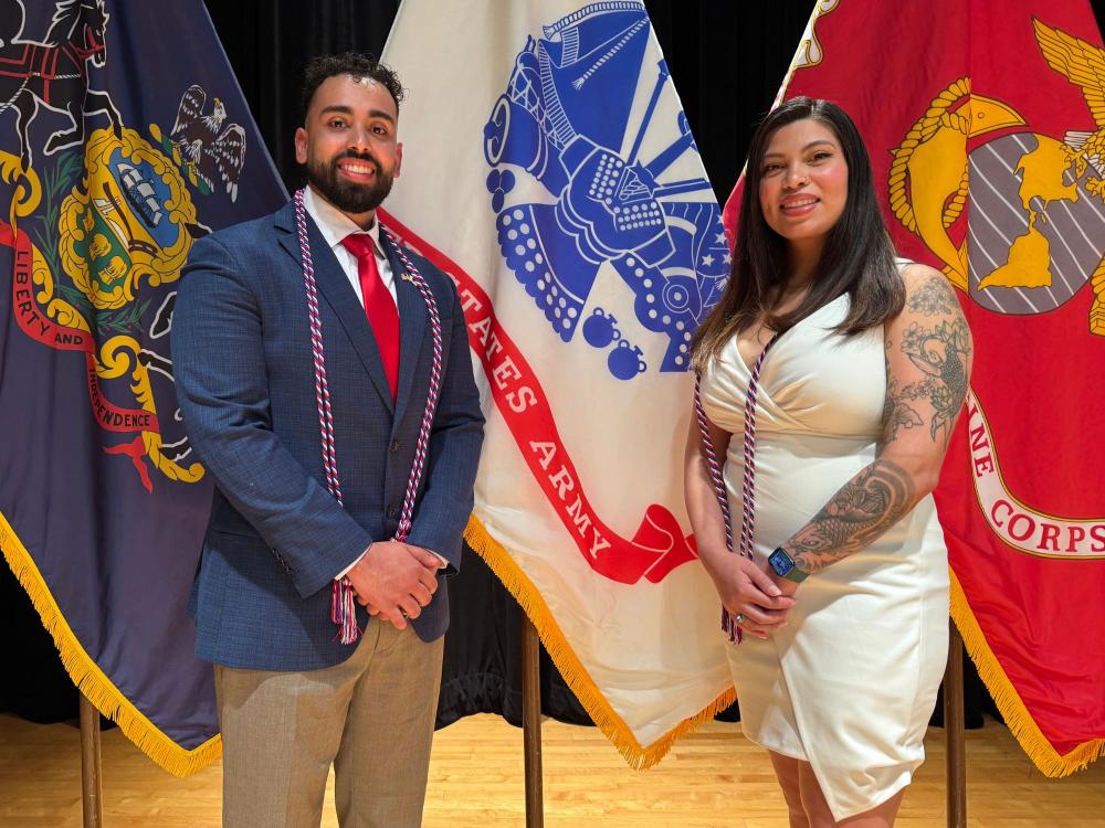 a man in a suit and woman in a dress stand in front of military flags