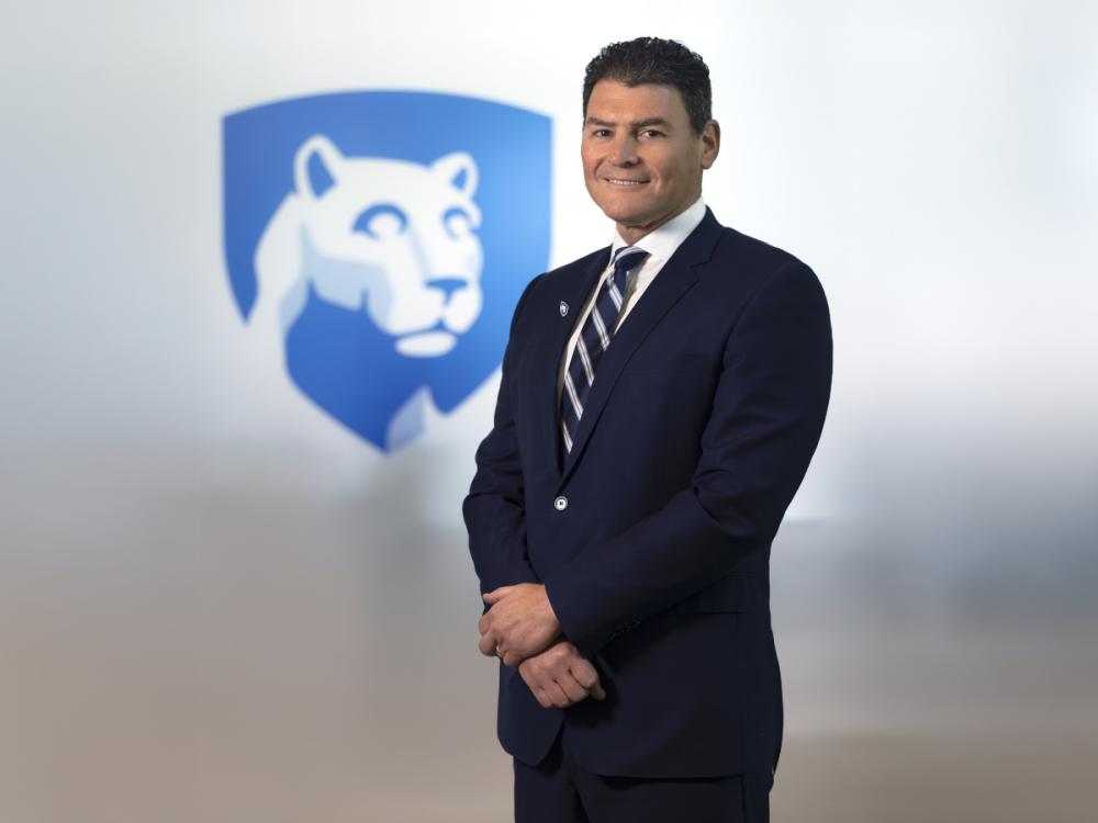 Michael Kupferman poses for a photo wearing a suit and tie. The Penn State Nittany Lion shield is in the background slightly out of focus.