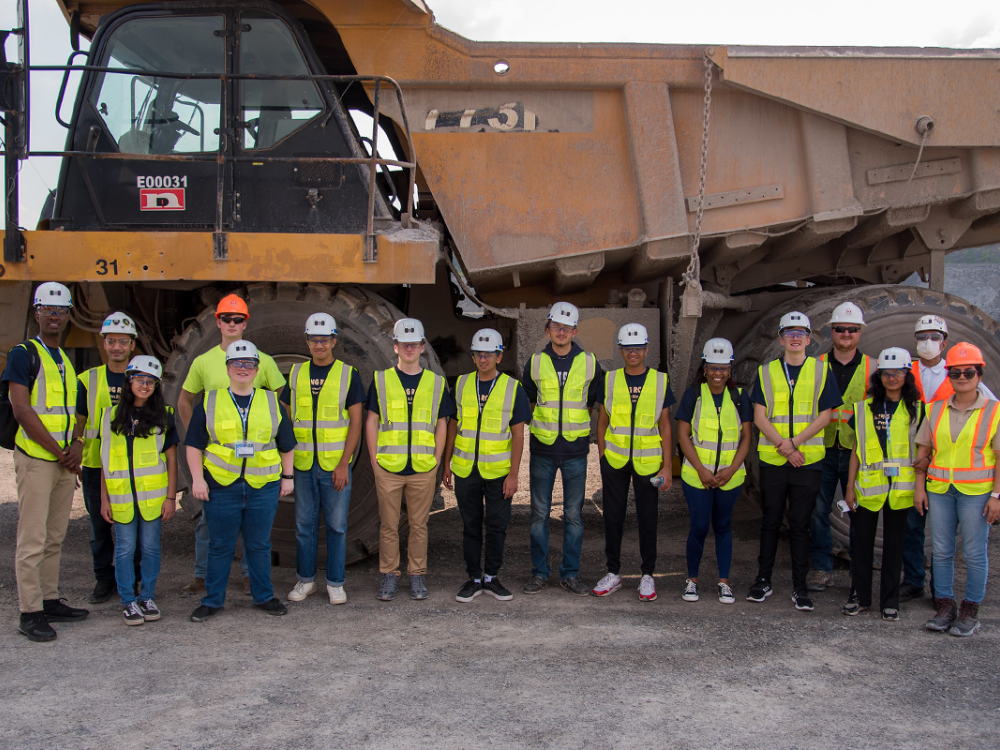 A group of students wearing safety helmets and high-visibility vests stand in front of a large yellow industrial truck at quarry.