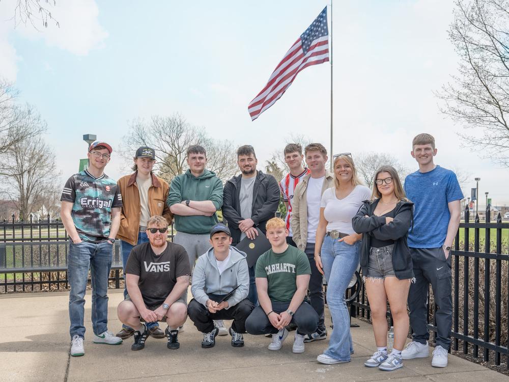 A group of students pose for a photo outside under an American Flag pole.