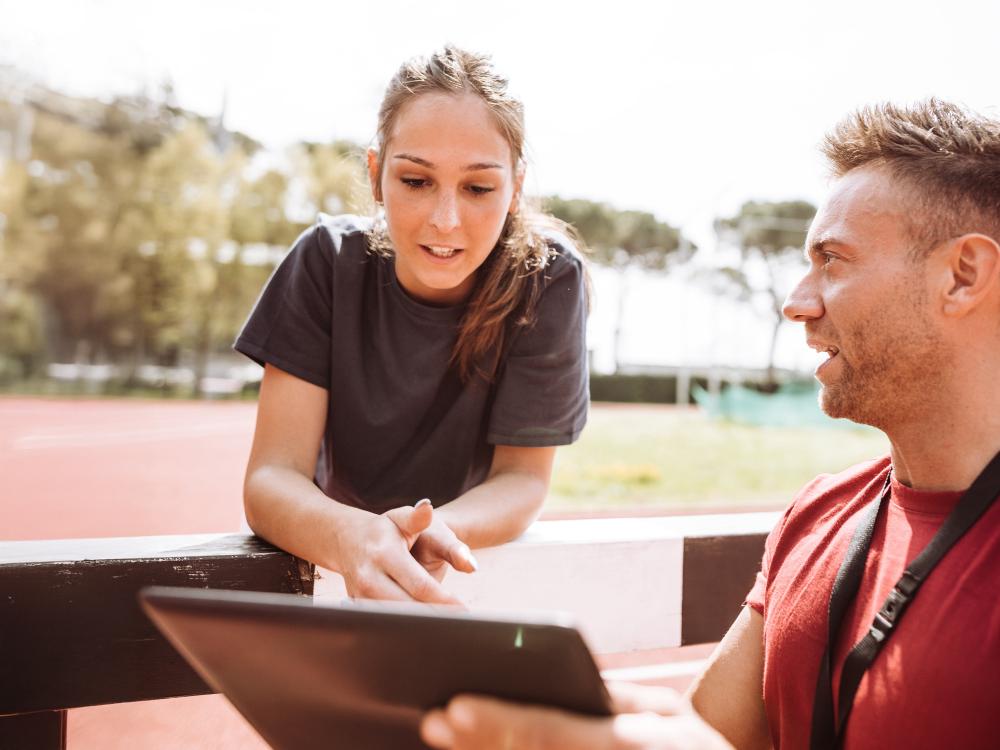 An athlete and coach, outdoors at a track, discuss something on a clipboard