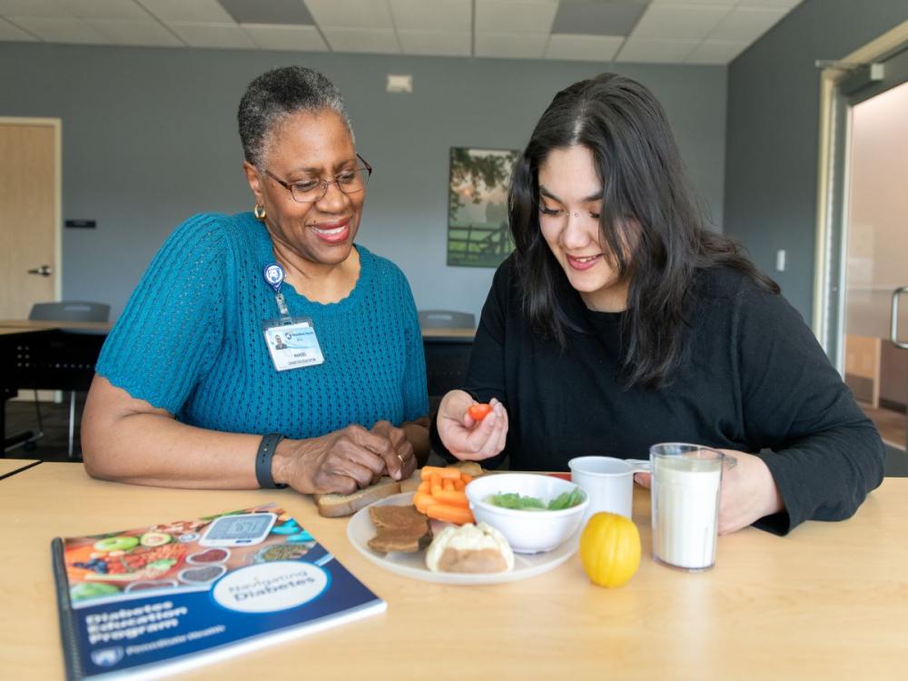 Two women sit at a table looking at a plate of food. A diabetes booklet is on the left. The woman on the left is wearing a knit top, glasses and a name badge. The woman on the right is wearing a long-sleeved top and is holding a cherry tomato.
