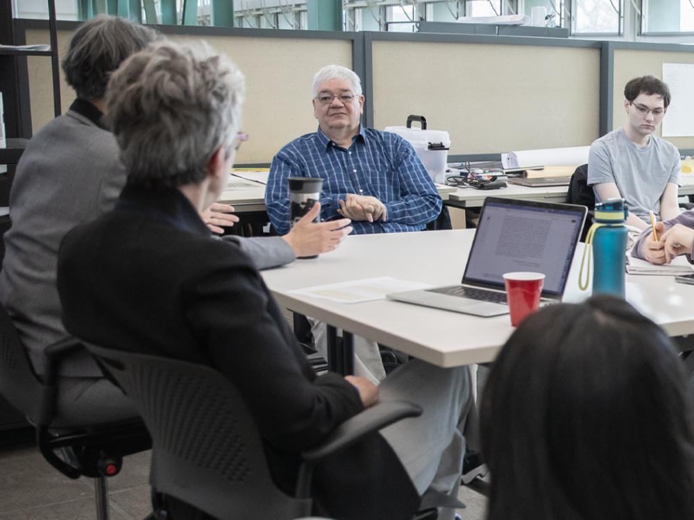 A man sitting at a table listening to someone on his right speak as others at the table look on.